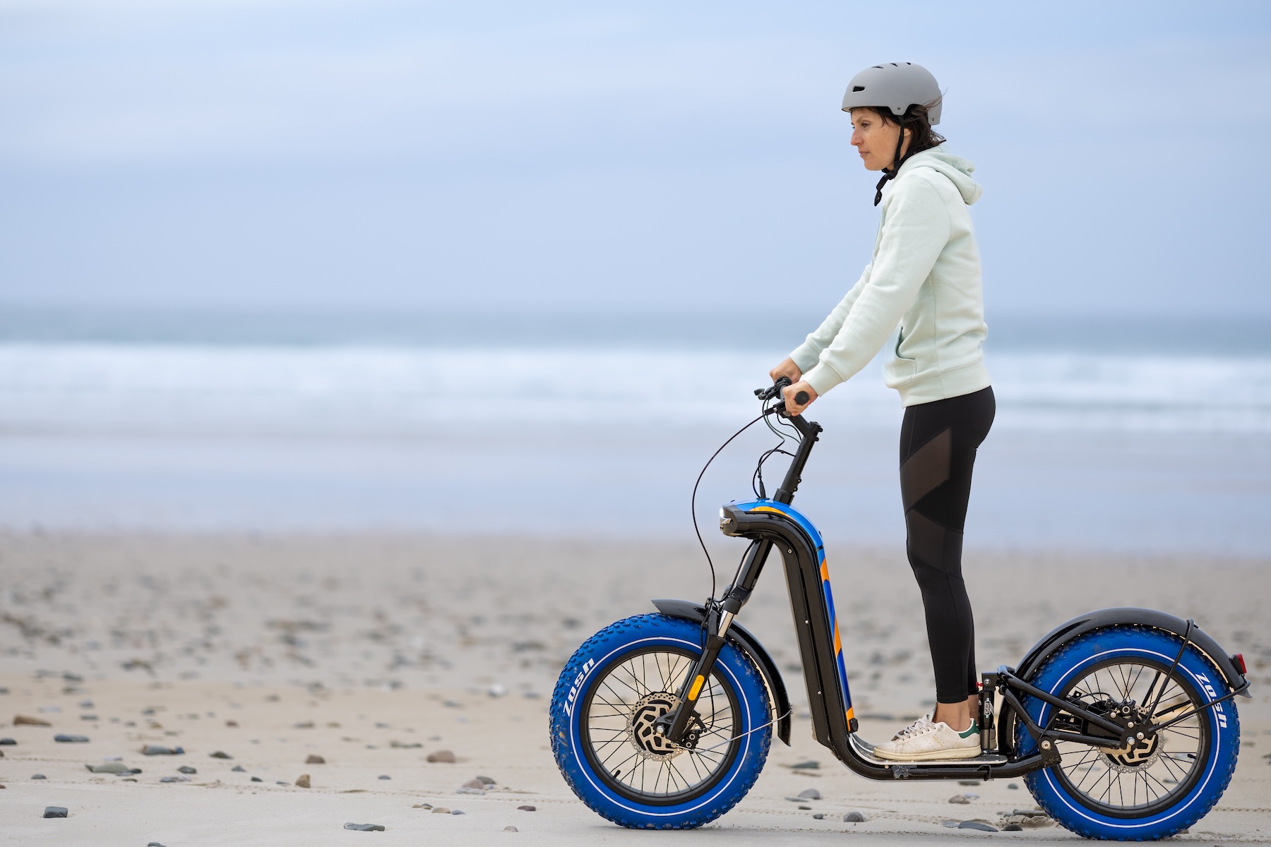Femme sur une trottinette électrique tout-terrain Zosh sur la plage de Biscarrosse, pneus larges bleus sur le sable