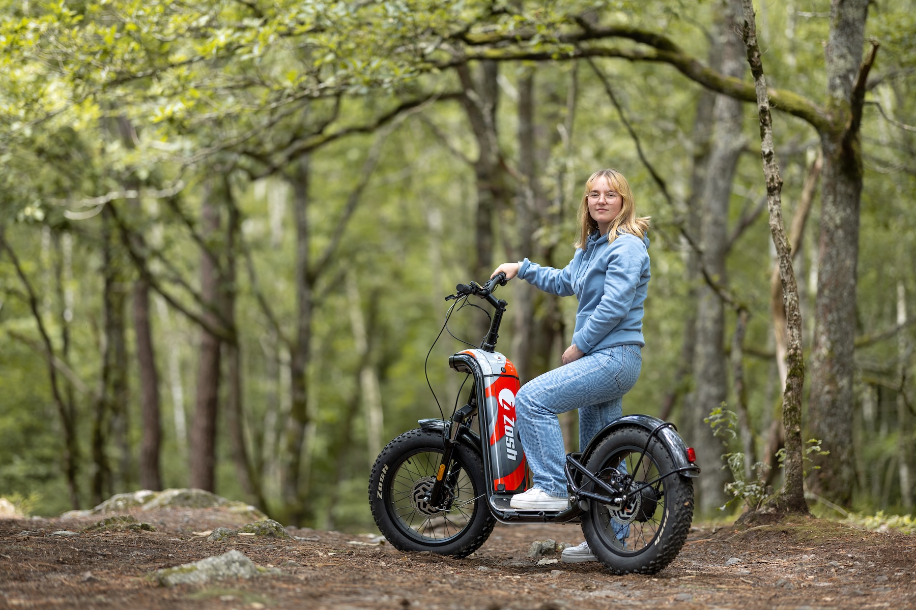 eune femme posant avec une trottinette électrique Zosh rouge sur un chemin forestier en sous-bois