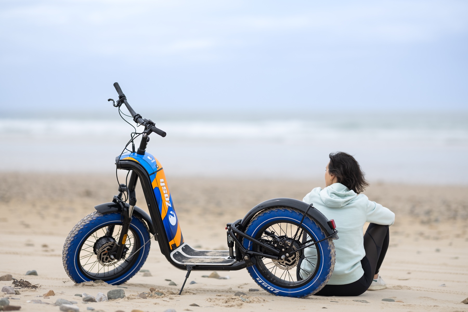 Femme assise sur le sable face à l'océan à côté de sa trottinette électrique Zosh bleu et orange