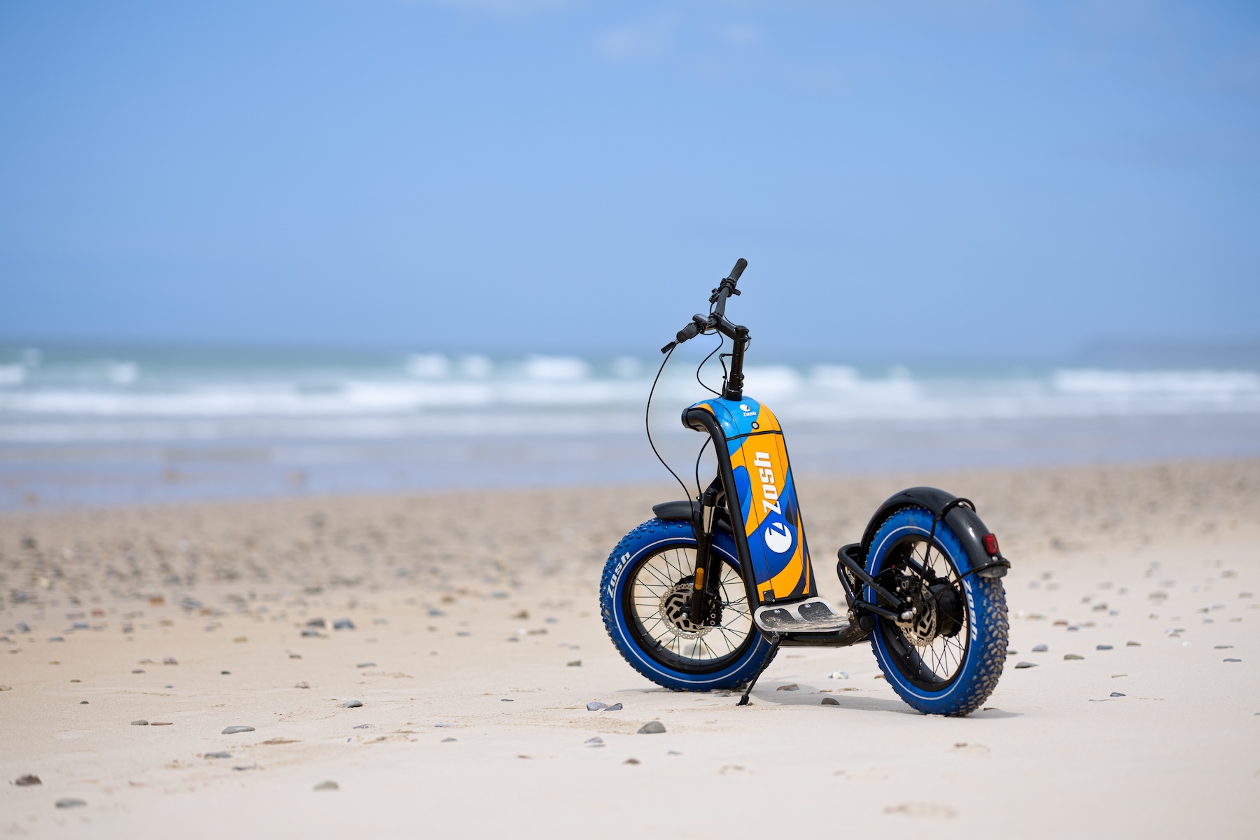 Trottinette électrique Zosh bleu et orange sur le sable avec les vagues de l'Atlantique en arrière-plan